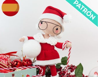 May include: A crocheted Santa Claus doll wearing a red and white suit, a white hat with a white pom-pom, and black shoes. The doll is holding a candy cane in one hand and a white ball in the other. The doll is surrounded by Christmas decorations, including candy canes, ornaments, and a box with a Christmas design.
