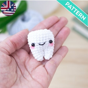 Puede incluir: Un diente de crochet blanco con una cara sonriente, mejillas rosadas y ojos negros. El diente está siendo sostenido en una mano.