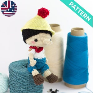 May include: Crochet pattern for a Pinocchio doll with a red pom-pom hat, blue pants, and a white shirt. The doll is sitting on a blue yarn ball.