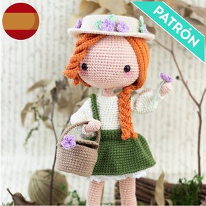May include: Crocheted doll with red hair wearing a white dress, green skirt, and a white hat with purple flowers. The doll is holding a brown basket with a purple flower and a single purple flower in her other hand. The doll is standing on a wooden surface with a green plant in the background.