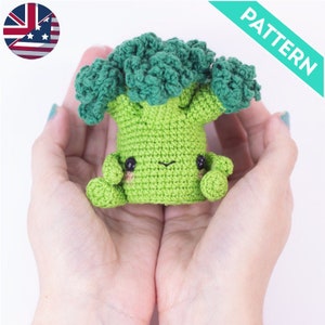 Puede incluir: Un juguete de peluche de brócoli de crochet verde con una cara sonriente y pequeños ojos negros. El juguete está siendo sostenido en las palmas de dos manos.