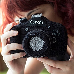 May include: A black crocheted camera with a gray lens and the text "Canon EOS 1100D" on the front.