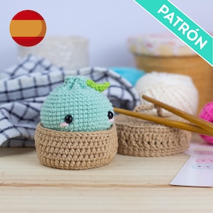 May include: A small, round, light green crocheted dumpling toy with black bead eyes and pink stitched cheeks, sitting inside a tan crocheted basket. Two wooden knitting needles rest nearby, and the word PATRÓN is visible in the corner.