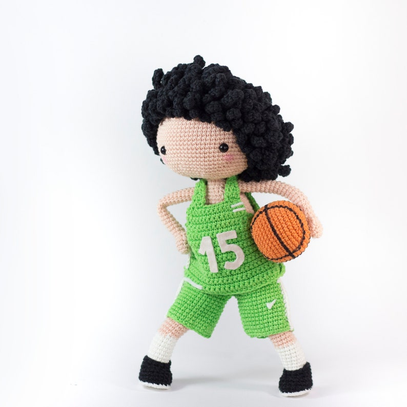May include: A crocheted doll wearing a green basketball uniform with the number 15 on the front. The doll is holding an orange basketball.