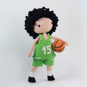 May include: A crocheted doll wearing a green basketball jersey with the number 15 on the front, black shorts, and white socks. The doll is holding an orange basketball.