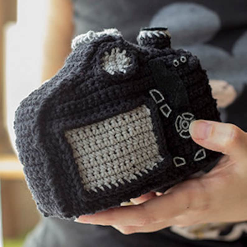 May include: A black crocheted camera with gray details. The camera has a lens, a flash, and buttons.
