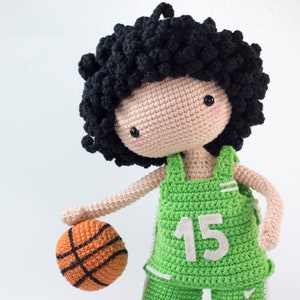 May include: Crocheted doll wearing a green basketball jersey with the number 15 on the front. The doll has black curly hair and is holding an orange basketball.