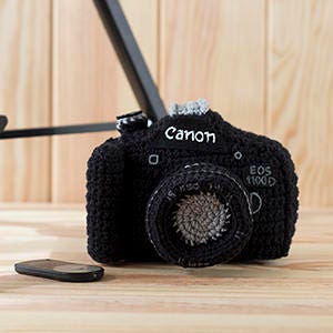 May include: Black crocheted camera with the word "Canon" and "EOS 1100D" embroidered on it.