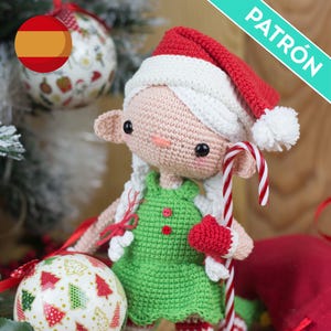 Op de afbeelding: Een gehaakte kerstelfpop met een rode en witte kerstmuts, groene jurk en wit haar. De pop houdt een zuurstok vast en heeft een rood hartvormig accessoire. Het woord "PATRÓN" is zichtbaar.