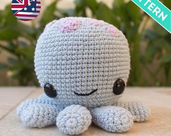 May include: A crocheted light blue octopus with pink accents on its head and black eyes. The octopus is lying on a light gray surface.