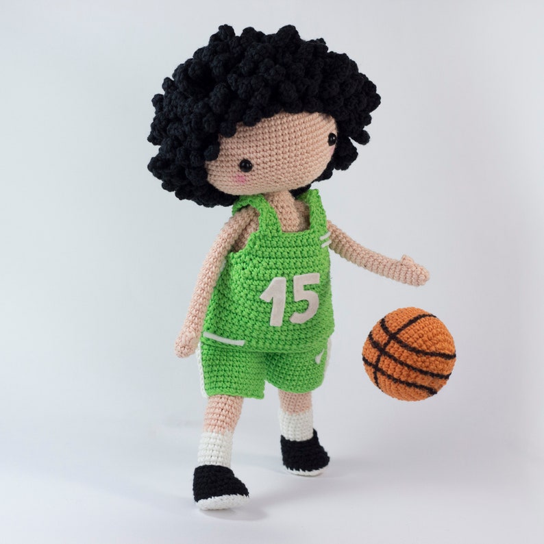 May include: A crocheted doll wearing a green basketball uniform with the number 15 on the front. The doll has black curly hair and is holding an orange basketball.
