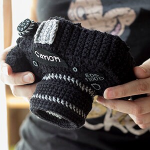 May include: A black crocheted camera with gray details. The camera has the word "Canon" written on the front and "EOS 1100D" on the side.