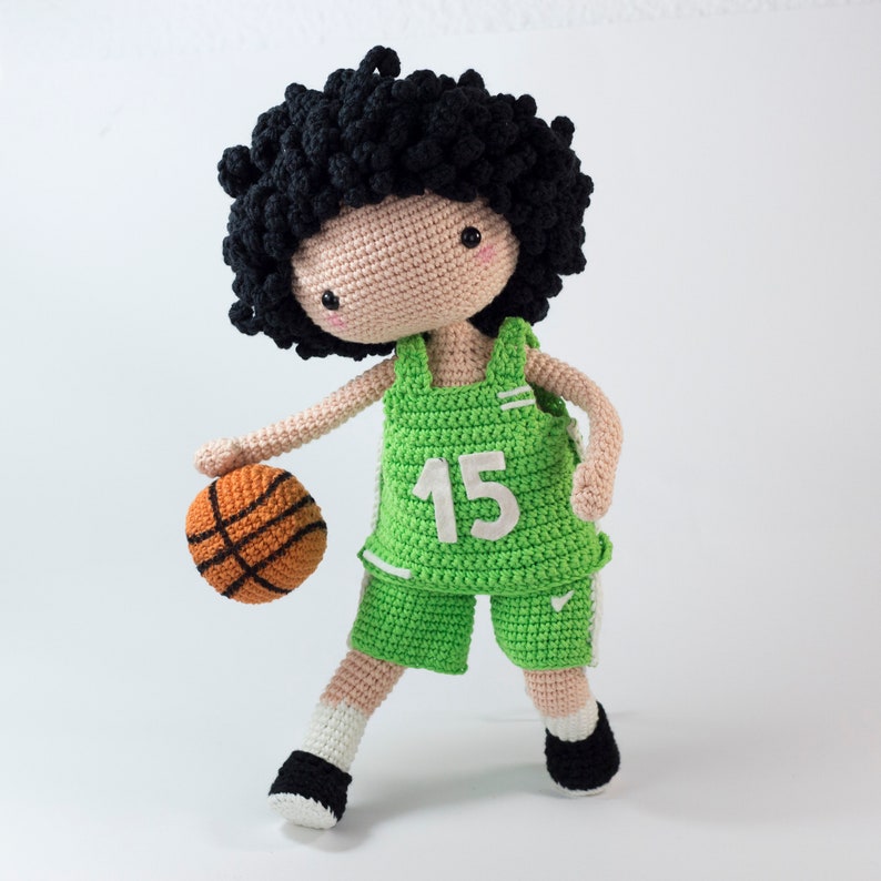 May include: A crocheted doll wearing a green basketball uniform with the number 15 on the front. The doll is holding a basketball.