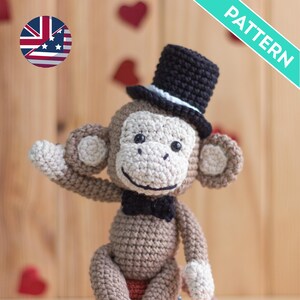 May include: A crocheted monkey wearing a black top hat with a white band and a black bow tie. The monkey is sitting on a wooden surface with red hearts in the background.