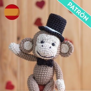 Könnte beinhalten: Ein gehäkelter Affe, der einen schwarzen Zylinder und eine schwarze Fliege trägt. Der Affe sitzt auf einer Holzoberfläche mit roten Herzen im Hintergrund.