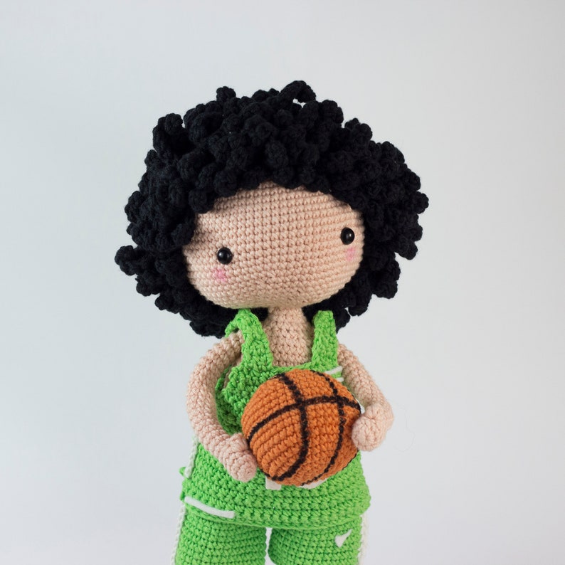 May include: A crocheted doll with black curly hair wearing a green and white basketball uniform and holding an orange basketball.