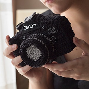 May include: A black crocheted camera with the brand name "Canon" and the model "EOS 1100D" written on it.