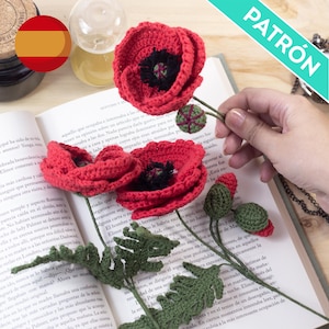 May include: Handmade red crochet poppies with black centers and green stems and leaves are displayed on an open book. The flowers are a vibrant red, and the leaves are a deep green. The word "PATRÓN" is visible in the image.
