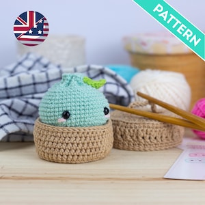 Puede incluir: Un patrón de ganchillo para un pequeño personaje de masa de color verde claro con ojos negros y mejillas rosadas, dentro de una cesta tejida de color canela. El set incluye dos ganchillos de madera y una tarjeta de instrucciones blanca.