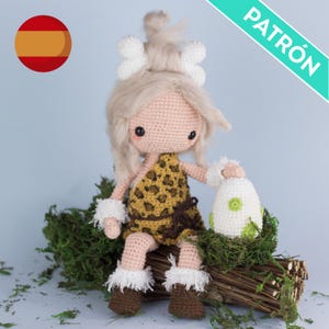以下が含まれることがあります： レオパード柄のドレスを着た洞窟の女性の、かぎ針編みアミグルミ人形のパターンです。人形は、苔と小枝のある丸太に座っています。パターンはスペイン語で入手できます。