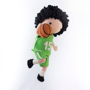 May include: A crocheted toy basketball player with black curly hair, wearing a green jersey with the number 15, and holding an orange basketball.