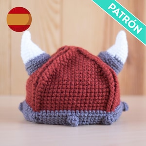 May include: A red and gray crocheted Viking helmet with white horns. The helmet is on a wooden surface. The word "PATRÓN" is written in white on a teal background.