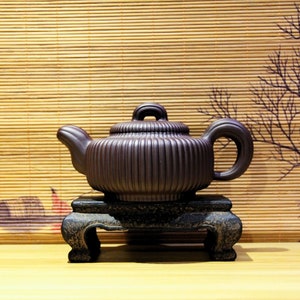 May include: A dark brown teapot with a ribbed design sits on a black wooden stand. The teapot has a rounded body and a spout on the side. The stand has three legs and a flat top.