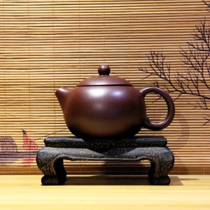 Peut inclure: Une théière en terre cuite foncée avec un corps rond et un petit couvercle est posée sur un support en bois noir. La théière est sur un fond de natte en bambou.