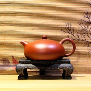 以下が含まれることがあります： 丸みを帯びたボディ、湾曲したハンドル、注ぎ口を備えた赤茶色のセラミックティーポット。 ティーポットは、暗い灰色の装飾的なスタンドに置かれています。 背景には竹のブラインドと装飾的な枝があります。