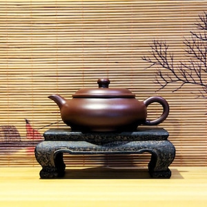 Op de afbeelding: Een donkerbruine theepot van keramiek met een deksel staat op een zwarte houten standaard met drie poten. De theepot heeft een rond lichaam en een tuit aan de zijkant. De standaard staat op een lichtbruin houten oppervlak.