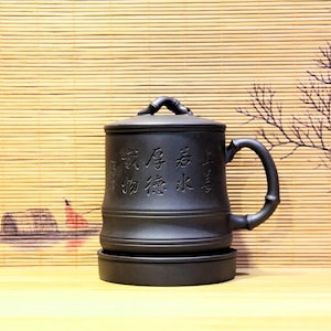 May include: A black ceramic tea mug with a lid and a bamboo-style handle. The mug has Chinese characters etched into the side and sits on a matching saucer. The background features a bamboo blind and a decorative branch.