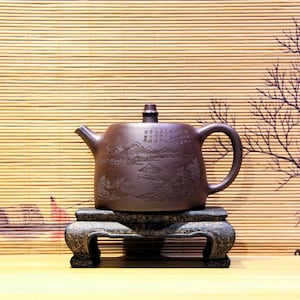 May include: A brown ceramic teapot with a landscape design and calligraphy, resting on a decorative stone stand. The teapot has a curved handle, a spout, and a lid. The stand has a carved design.