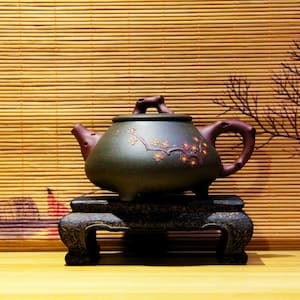 以下が含まれることがあります： 濃い緑色の茶器に、茶色の取っ手と花柄が施されています。茶器は、3本の脚が付いた暗い木製の台の上に置かれています。