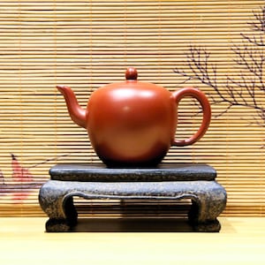 May include: A reddish-brown ceramic teapot with a rounded body, curved spout, and loop handle. The teapot sits on a dark, ornate, rectangular stand. The background features a bamboo blind and a decorative branch.
