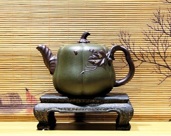 May include: A green ceramic teapot with a pumpkin-like shape, featuring a dark brown handle, spout, and lid. The teapot is decorated with a floral design and sits on a dark, ornate stand. The background includes a bamboo blind.