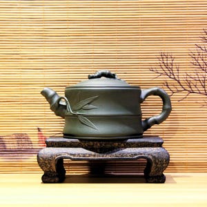Op de afbeelding: Een donkergroene keramische theepot met een bamboe-stijl handvat en tuit, rustend op een decoratieve, donkergekleurde standaard. De theepot heeft bamboeblad details en een bamboe-vormige dekselgreep.