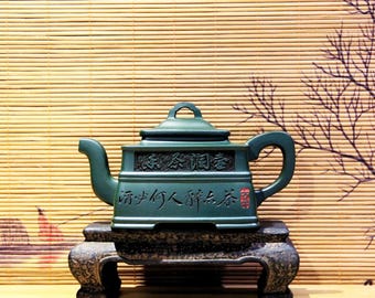 以下が含まれることがあります： 複雑な彫刻と中国語の文字が刻まれた濃い緑色の陶器製の急須が、濃い木製の台の上に置かれています。急須には蓋と注ぎ口があり、台には3本の脚があります。