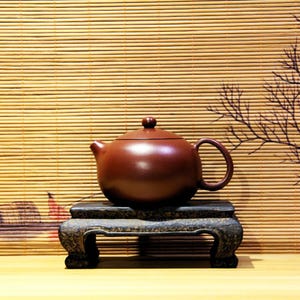 May include: A dark brown ceramic teapot with a rounded body and a lid, sitting on a black wooden stand with three legs. The teapot is on a light brown wooden surface.
