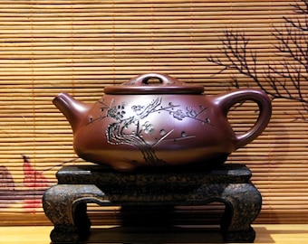 May include: A brown ceramic teapot with a floral design sits on a black wooden stand. The teapot has a lid with a small knob on top. The stand has three legs.