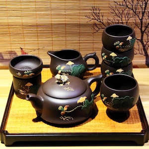 May include: A brown tea set with a teapot, a pitcher, and four cups. The teapot has a frog on the lid and a floral design. The cups and pitcher have a similar floral design. The set is on a wooden tray.