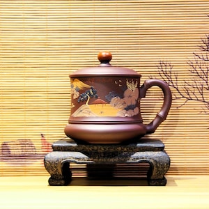 May include: A brown ceramic teacup with a lid and a handle. The teacup has a painted design of a landscape with mountains, trees, and a house. The teacup is sitting on a wooden stand.
