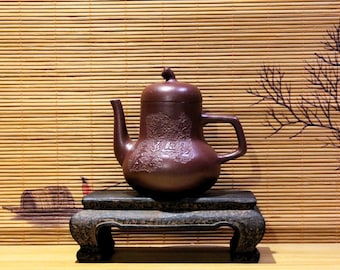 May include: A dark brown teapot with a carved design sits on a black wooden stand. The teapot has a rounded body and a spout on the left side. The stand has three legs and is made of wood.