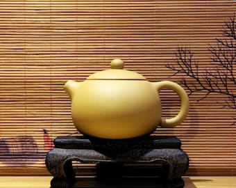 May include: A light brown ceramic teapot with a rounded body and a small spout. The teapot is sitting on a dark wooden stand with a simple design.