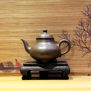 Puede incluir: Una tetera de cerámica marrón con tapa, boquilla y asa, colocada sobre un soporte oscuro y ornamentado. La tetera presenta un diseño de paisaje detallado. El fondo incluye una persiana de bambú y una rama de árbol decorativa.