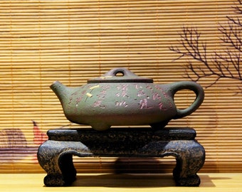 May include: A dark green teapot with a lid and a handle, decorated with gold and pink Chinese characters. The teapot is sitting on a black wooden stand with three legs.