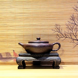 May include: A brown ceramic teapot with a curved spout and handle, resting on a dark, ornate stand. The teapot has a lid and is decorated with floral patterns. The background features a bamboo blind and a branch silhouette.