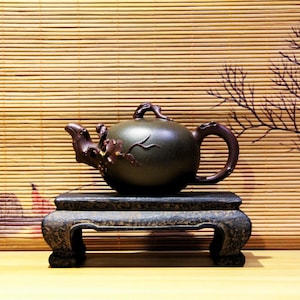 以下が含まれることがあります： 茶色の取っ手と注ぎ口が付いた、濃い緑色の陶器製の急須。枝と葉の浮き彫りの模様で飾られています。急須は、3本の脚が付いた濃い茶色の木製台の上に置かれています。