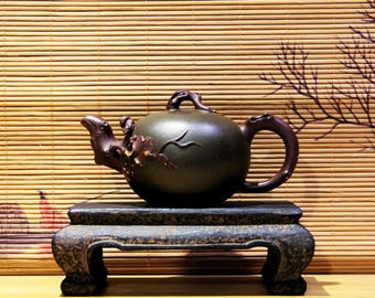 May include: A dark green ceramic teapot with a brown handle and spout, decorated with a raised design of branches and leaves. The teapot is sitting on a dark brown wooden stand with three legs.