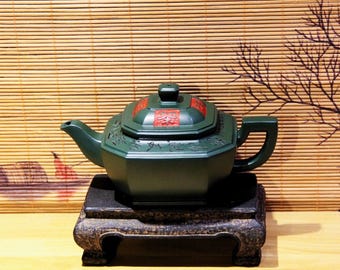 May include: A dark green teapot with a square body and a lid with red accents. The teapot is sitting on a black wooden stand with three legs.