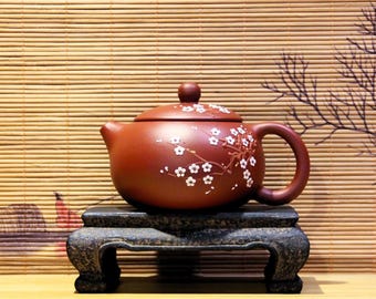 May include: A red clay teapot with a white floral design sits on a black wooden stand. The teapot has a lid and a spout. The stand has three legs.